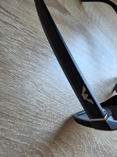 The image shows a close-up, angled view of a pair of black sunglasses resting on a light brown wooden surface. The sunglasses are partially visible, with the arm and a portion of the frame in focus. A logo, appearing as stylized white or silver lettering, is embossed on the exterior of the sunglasses arm. The wooden surface has a visible grain pattern running diagonally across the frame. There are no people or actions depicted in the image. The location in Mijas, Spain is not discernible from the visual information provided.