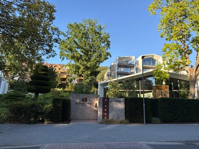 The image depicts the entrance to the Alea Resort in Bad Orb, Germany. The entrance is marked by two large stone pillars. The left pillar displays the resort's logo and name, "ALEA RESORT BAD ORB". The right pillar features several smaller signs with names like "FOGO," "BEAUTY AND SOUL," "MUERNIST," and "REVEL HOUSE," along with a number "1" and a building number "-A5-49-".

Behind the entrance, a modern building with glass facades and white architectural elements is visible. The building is surrounded by lush greenery, including manicured bushes, trees, and a topiary. The sky is clear and blue, suggesting a sunny day. A paved walkway with interlocking gray bricks leads towards the entrance. A streetlight stands on the right side of the walkway in front of the building. The overall scene conveys a sense of upscale and tranquil accommodation.
