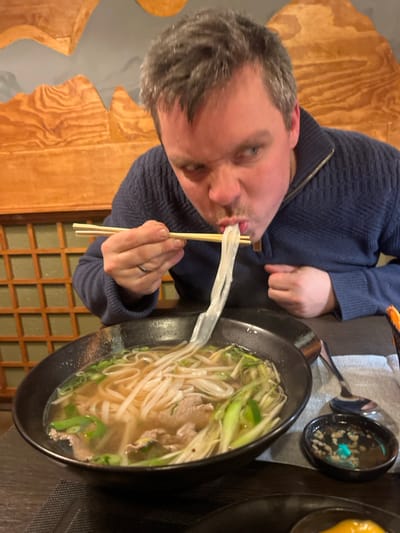 A person with graying hair and a blue sweater is eating a long white noodle using chopsticks. The person is looking at the camera with a slightly skewed expression. The noodle stretches from the chopsticks directly into the person's mouth. In front of the person is a large black bowl filled with broth, rice noodles, bean sprouts, and green onions, consistent with Pho soup. A small dish with a dark sauce is visible near the soup, and the background has light brown wood paneling.
