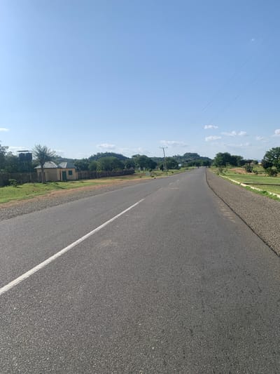 An asphalt road stretches into the distance under a clear blue sky dotted with a few white clouds. A white line marks the left lane of the road. On the left side of the road, a concrete wall encloses a property with two small, light-colored buildings with green doors and windows, and two water tanks on a raised platform. Beyond the wall, green grass and trees are visible, with rolling hills covered in vegetation in the background. On the right side of the road, the shoulder is covered in gravel, and beyond that, there are grassy fields and scattered trees. Utility poles and power lines are present along the right side of the road. The scene appears to be in a rural or semi-rural area of Boi A, Nigeria, given the infrastructure and landscape. There are no people clearly visible in the image.
