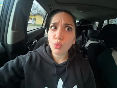 An indoor photograph, likely captured from the interior of a vehicle, depicts a young adult female with dark hair and light skin, positioned in what appears to be the front passenger seat. The female subject exhibits a facial expression with wide-open eyes rolled upward and to the right, and her mouth is pursed into a "fish lips" shape. She is wearing a black hooded sweatshirt featuring a white trefoil logo, partially visible on the front. The vehicle's interior consists of black upholstery; a black headrest is visible to the subject's right, and the rear seats contain various items, including a dark child car seat with visible "RECARO" branding and additional bags. Through the passenger-side window, an outdoor scene in Lonate Pozzolo, Italy, is visible, featuring a yellow commercial building with a roof overhang. A white BMW sedan is parked outside this structure. Bare trees and an overcast sky indicate a non-summer season and dim lighting conditions.