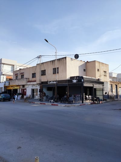 A street scene captured in Tunisia, likely during the late afternoon or early evening as suggested by the low angle of the sun and the shadows cast. The weather appears clear and pleasant, with a bright blue sky overhead and minimal cloud cover.
The primary subject is a building at a street corner, housing a cafe named "Caffein," which is clearly visible on a black awning above the entrance. The cafe has outdoor seating, with several tables and chairs arranged on the sidewalk, some occupied by patrons. The building itself is a light-colored, multi-story structure with balconies and windows characteristic of North African architecture. Adjacent to the cafe are other businesses, including one with signage that reads "LOUIATIPNEUS" and another with Arabic script.
In the foreground, a paved street is visible, showing signs of wear and tear. On the left side of the street, a small, light-colored car is parked. Several people are present in the scene, including individuals walking on the sidewalk and sitting at the cafe. One person in the foreground appears to be a woman walking with a child.
Notable details include a street lamp extending over the sidewalk, power lines crisscrossing overhead, and a satellite dish mounted on the building. The overall atmosphere appears relaxed and everyday, reflecting a typical street scene in a Tunisian town or city.