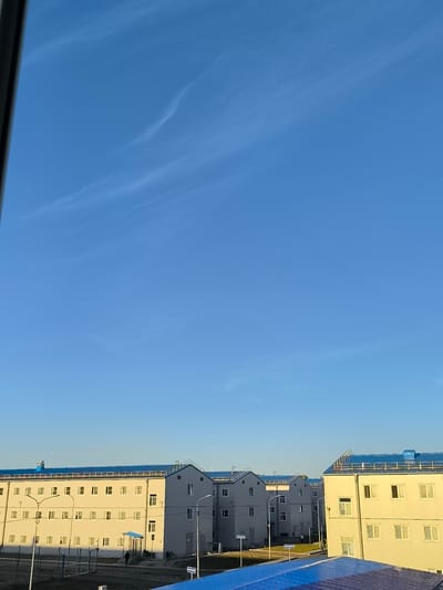 The image depicts a scene in Ust-Luga, Russia, featuring a row of identical, modern, light-colored buildings with blue roofs. These multi-story structures appear to be modular or prefabricated housing units. The buildings are organized in a grid-like fashion, suggesting a planned development. Several streetlights are visible between the buildings. The sky is clear and blue with faint wispy clouds. In the foreground, a fence separates the building area from a dirt or gravel path. A few individuals are present on the path, walking or standing. One building in the foreground is brightly lit by sunlight, highlighting its facade and numerous windows. The overall impression is one of new construction or a recently completed development in a somewhat industrial or developing area.