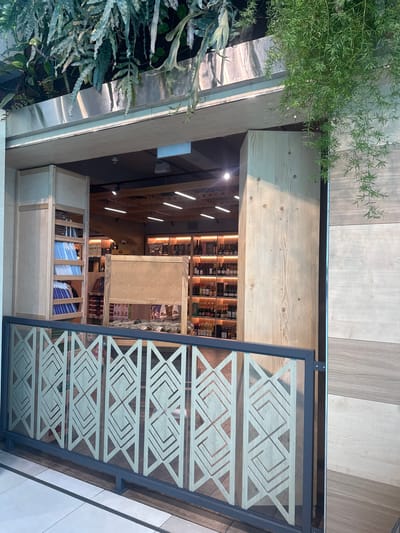 The image shows the entrance to a store in Bubuieci, Moldova. The store has a natural wood facade and is decorated with hanging plants. In the foreground, there is a metal railing with an abstract geometric pattern. Inside the store, shelves are stocked with various products, including bottles of wine and other items. The lighting suggests it is daytime. There are no people visible in the image.