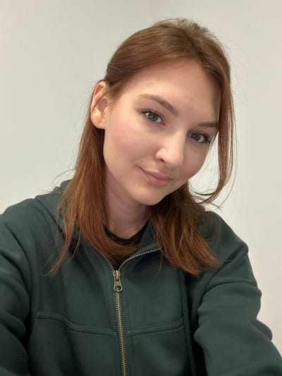 The media file shows a close-up, portrait-style photograph of a young woman. She has auburn hair that is styled in loose waves around her face. She is wearing a dark green hooded jacket with a zipped front. The background is a plain, light-colored wall, suggesting an indoor setting. The lighting is soft and even, indicating it could be daylight or well-lit indoor conditions, with no strong shadows. The woman is looking towards the camera with a gentle, almost playful expression. Her gaze is directed slightly off-camera, and her lips are parted slightly as if she is about to speak or has just finished. The image is oriented sideways, with the top of her head towards the left. There is no visible text within the image. The overall impression is casual and personal.
