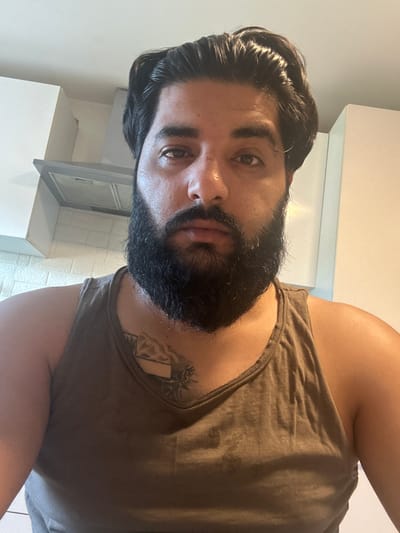 A selfie taken from a slightly low angle captures a man with dark hair and a full beard. He is wearing a dark brown tank top, revealing a tattoo on his chest. The background appears to be an indoor kitchen area with white cabinets and a stainless steel range hood visible. The lighting suggests it is daytime, with natural light entering the space. The man's gaze is directed towards the camera. The specific location in the city of Mainburg, Germany, is not determinable from the provided image.