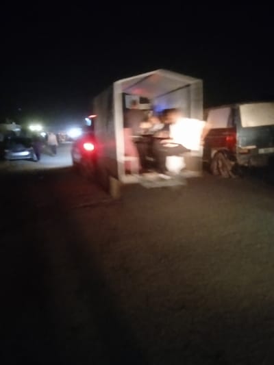 This is a nighttime image captured in Al-Mawasi, Palestinian Territories, depicting an outdoor scene characterized by low light and motion blur. The foreground shows a dark, possibly unpaved ground surface. To the right, an open-sided, canopy-like structure, illuminated from within by bright artificial light, stands on the roadside. Inside this structure, several indistinct human figures are visible, appearing engaged in activity, with one individual wearing a light-colored top. To the left of this structure, a vehicle with a prominent red taillight is present, facing away from the viewer. Further to the right, behind the illuminated structure, another vehicle, possibly a dark van or truck, is partially visible. In the distant left background, multiple bright artificial light sources, likely from vehicles or streetlights, illuminate a section of what appears to be a road or open area, where several other indistinct human figures can be observed. The overall environment is dark, with artificial lighting being the primary source of illumination.