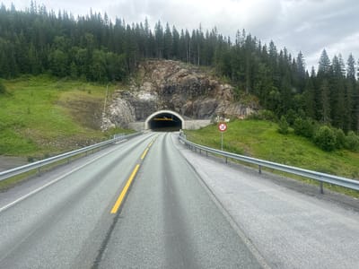 This media file depicts a scenic view of a road approaching a tunnel entrance in Mosjøen, Norway. The road is paved with asphalt and marked with yellow and white lines. On either side of the road are metal guardrails. The surrounding landscape is characterized by a lush, green hillside covered in grass and trees, with a dense forest of pine trees covering the upper slopes.
The tunnel entrance is carved into a rocky mountainside. The tunnel itself is arched and illuminated, with a yellow and black striped height restriction sign visible above the entrance. There is also a traffic sign on the right side of the road, a red circle with a diagonal line through it, indicating a prohibition. The sky is overcast with clouds, suggesting a possibly cool or damp day, typical of Norwegian weather. The time of day appears to be daytime, but the cloudy conditions create a diffused light.
There are no people visible in the frame, and the focus is on the natural environment and the man-made infrastructure of the road and tunnel. The overall impression is one of a journey through a natural, somewhat wild landscape, with the tunnel representing passage through the mountain. The image conveys a sense of tranquility and the vastness of nature.