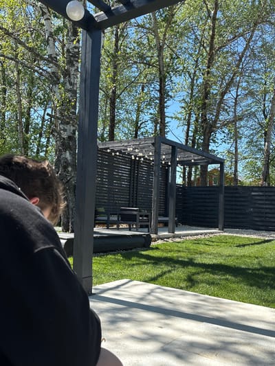 The image depicts an outdoor scene, possibly a backyard or garden in Юг, Russia. A black wooden structure with string lights is visible, situated on a stone path adjacent to a grassy area. Behind the structure is a dark wooden fence, and trees with light green foliage fill the background under a clear sky. A person is partially visible in the foreground, wearing a black garment. Their head and shoulder are visible, suggesting they are close to the camera.