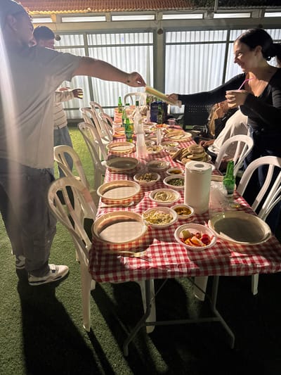 This image depicts an outdoor evening gathering at a residence or event space in Kfar Yona, Israel. The scene is illuminated by overhead artificial lighting.
A long rectangular folding table, covered with a red and white checkered tablecloth, serves as the central focus, laden with numerous food and beverage items. Surrounding the table are multiple white plastic chairs. The ground beneath is covered with artificial green turf.
In the foreground left, a man wearing a light-colored short-sleeved t-shirt, dark cargo pants, and white sneakers is extending his right arm to hand a bundle of colorful drinking straws to a woman. A small tattoo is visible on his right forearm. Behind him, another man in a striped long-sleeved shirt is partially visible.
In the mid-ground right, a woman with dark hair pulled back, wearing a dark long-sleeved top, smiles as she reaches out with her left hand to receive the straws. She holds a transparent cup with a pink straw in her right hand. Part of another seated person is visible behind her.
The table features a variety of dishes: numerous small white plastic bowls contain various dips and salads, including what appears to be hummus and other mixed greens or legumes. Several larger beige plastic plates, some stacked, are set out, along with a stack of small clear plastic cups. A roll of white paper towels stands upright on the table. A pile of pita bread or similar flatbread is present. Beverages include several green glass bottles, some identifiable as Carlsberg beer, and other dark-labeled bottles. A small white bowl contains cut red and yellow fruit or vegetables. Gold-colored plastic forks are laid on the tablecloth.
The background reveals a covered patio or pergola structure with a tiled, reddish-brown roof and translucent or frosted panel walls, reflecting the bright artificial lights.