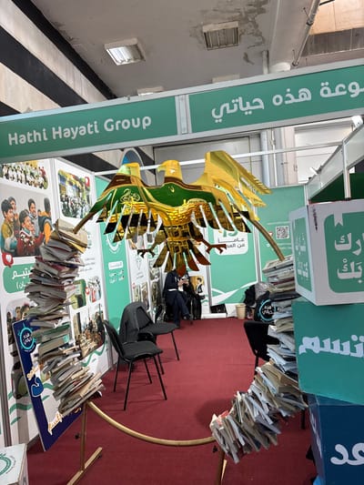 The scene depicts an indoor exhibition booth located in Al-Ghizlaniya, Syria. The floor is covered with a red carpet. Overhead, an industrial-style ceiling with black and white striped beams, visible conduits, and fluorescent light fixtures shows signs of wear and minor water damage.

The booth, prominently labeled "Hathi Hayati Group" and "مجموعة هذه حياتي" in Arabic on a large green header, is enclosed by light green panels. These panels feature photographic collages of children and adults, Arabic text, and a QR code linked to "hathi-hayati.com".

A large, stylized golden eagle sculpture, appearing reflective and multi-layered with green accents and Arabic calligraphy, is centrally positioned. Two black chairs are visible in the mid-ground, with one tipped forward. Two adults are seated behind the eagle sculpture; one is occupied with a mobile phone.

Book displays form significant elements of the booth: a tall, vertical stack of diverse books stands on the left, while a curvilinear arrangement of books forms an arc on the right, supported by a golden railing and culminating near two stacked boxes. The upper green box displays the Arabic text "شارك كتابك" (Share your book), and the lower blue box shows "و ابتسم" (and smile). A visible book cover in the left stack also bears the title "هذه حياتي" (Hathi Hayati).