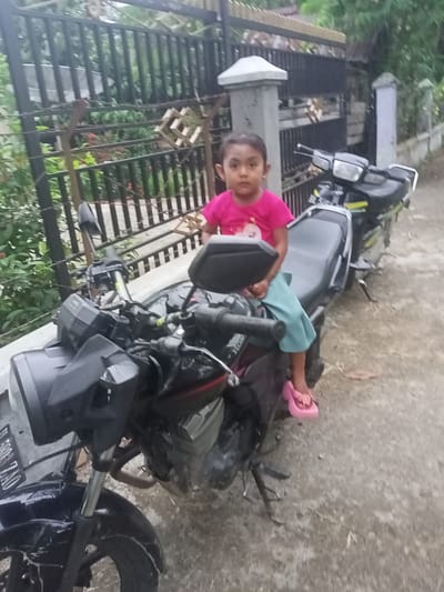 An outdoor scene in Teupinjok, Indonesia, depicts a young girl seated on a black motorcycle, with a second motorcycle parked behind it. The girl, approximately 3-5 years old, wears a pink t-shirt with a light graphic, light blue or teal pants, and pink flip-flops. Her dark hair is pulled back, and she looks directly at the camera with a neutral expression. The motorcycle in the foreground is black, featuring a rectangular headlight and a partially visible front license plate reading "F 5900 ZAO". The second motorcycle, further back and to the right, is black and green or yellow. Both motorcycles are stationary on their kickstands. The ground is a light-colored, textured path, possibly dirt or worn concrete. In the background, a dark metal fence and gate with concrete pillars separate the path from lush green vegetation and trees. A decorative wooden lattice structure hangs on the fence to the right. A light-colored curb or wall is visible between the motorcycles and the fence.