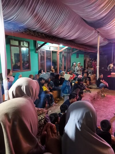 The image depicts a nighttime community gathering under a temporary canopy in Kecamatan Cimaung, Indonesia. A diverse group of people, comprising adults and children, are seated on the ground and on low concrete ledges, their attention focused towards an unseen point to the right of the frame.

In the foreground, the backs of two individuals wearing light-colored hijabs are prominent. The middle ground reveals numerous attendees, many of whom are women also wearing hijabs, alongside men and children. They are seated in a dense arrangement, observing the event unfolding.

The backdrop features a house with distinctly green-painted walls, traditional brown wooden window frames, and wooden doors. Artificial lighting, likely from overhead or strategically placed sources, illuminates the scene, creating a warm ambiance against the dark night. To the far right, a large speaker and other sound equipment are visible, suggesting that the gathering involves amplified sound, possibly for a performance, speech, or screening. The ground beneath the attendees appears to be a mix of dirt and uneven concrete, with some people utilizing raised platforms for seating. The atmosphere is one of communal engagement and quiet observation. No text is visible within the image.