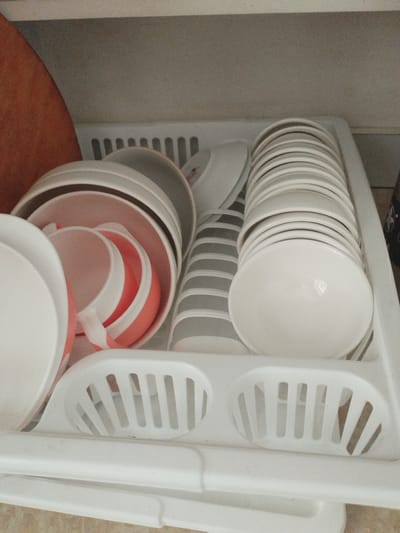 In Октябрь, Kyrgyzstan, a white plastic dish rack is filled with clean dishes. The rack is positioned on a countertop, likely in a kitchen. Among the dishes are several white plates stacked vertically, a set of pink and white striped cups, and some smaller white bowls. The dishes appear to be made of ceramic or porcelain. The environment is indoors, with the background showing a light-colored wall and a wooden object, possibly a cutting board, to the left. The lighting is diffused, suggesting it's not direct sunlight. There are no people visible in the image, and no actions are being performed. The scene is static and appears to be a typical domestic setting.