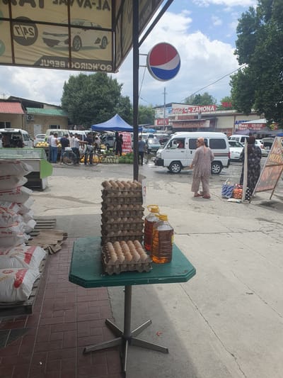 The image depicts a street market scene in Avval', Uzbekistan, under a partly cloudy sky. In the foreground, a green octagonal table displays stacks of egg cartons and two large bottles of amber liquid, likely cooking oil. To the left, sacks of "AVVAL DON" are piled. The market stalls and vehicles suggest a commercial area. In the background, people are engaged in various activities, such as shopping and conversing. Several vehicles, including vans and cars, are parked or moving through the market. Prominent signage includes a Pepsi logo, "ZAMONA," and advertisements for taxis. The overall environment appears to be a bustling outdoor marketplace.