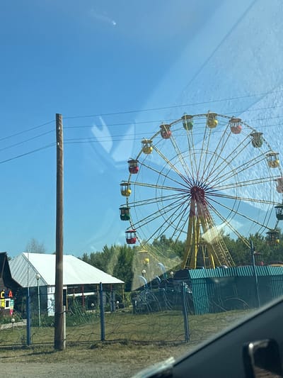 The image depicts an outdoor amusement park on a clear, sunny day. A large, colorful Ferris wheel, primarily yellow and red, dominates the right side of the frame, with multiple passenger cars visible. To the left of the Ferris wheel, a white-roofed building with a wooden facade is present, featuring a sign with the letters "CA" visible. A utility pole stands prominently in the foreground, with wires running across the scene. A decorative fence separates the immediate foreground from the amusement park area, which includes a green corrugated metal structure behind the fence and trees in the background. The image appears to be taken from a vehicle, as evidenced by reflections on the glass. The location is identified as Aya, Russia. There are no people or animals visible in the image.