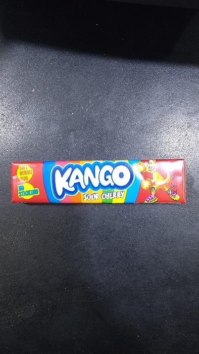The image features a single, rectangular packet of "KANGO" bubble gum, positioned centrally on a dark, textured surface. The gum wrapper is primarily red, with a prominent blue "KANGO" logo outlined in white. Vertically oriented yellow text on a green background to the right of the logo identifies the flavor as "SOUR CHERRY."
The top portion of the wrapper showcases colorful, cartoon-style illustrations of what appear to be kangaroos or similar lively figures in various poses, rendered in shades of orange, green, and purple, against a red and blue background. At the bottom-left of the packet, two yellow circular seals are visible. One clearly states "SOFT BUBBLE GUM," while the other, partially obscured, indicates "NO STICKING," implying a non-stick formula.
The background surface is dark, appearing to be a countertop or table with a slightly grainy or speckled texture, and shows some minor dust or debris. Lighting from the right illuminates parts of the surface and the gum packet, creating a slight sheen. There are no other discernible objects or specific environmental details in the image that would visually confirm the location as Yakkatut, Uzbekistan; it is a straightforward product photograph.