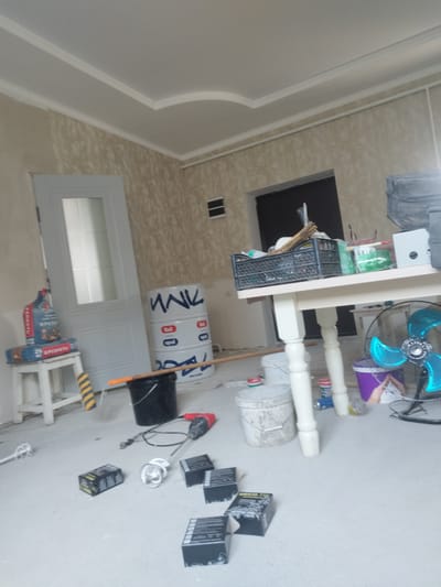 The image depicts an indoor construction or renovation scene. The room is unfinished, with plain walls and ceiling. Various tools and materials are scattered around, suggesting ongoing work. In the foreground, there are several black boxes, possibly containing hardware or supplies, and a power tool, likely a mixer or drill. To the left, a white stool holds two bags of what appears to be building material, and a paint roller with a yellow and black striped handle rests nearby. Behind the stool is a white door with glass panels. To the right, a white table is laden with more items, including a black plastic crate filled with tools or supplies, and some green objects that could be paint cans or sealant. A large white barrel with blue lettering is situated behind the stool, and a black bucket is on the floor next to it. Further to the right, a blue electric fan is positioned near the table. In the background, there's a dark doorway. The floor is concrete and appears dusty. The overall impression is one of a workspace that is actively being used for construction. The scene is located in Tokmok City, Kyrgyzstan.