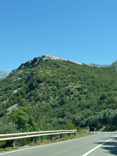 The image depicts a daytime scene in Blizikuće, Montenegro, showcasing a mountainous landscape with a road in the foreground. The upper third of the image is dominated by a clear, bright blue sky. A verdant, tree-covered mountain slope occupies the mid-ground and background, exhibiting rocky outcrops, particularly on the left side. Perched along the ridge of this mountain is a settlement composed of multiple white-walled buildings with distinctive red-tiled roofs. A prominent white structure, resembling a church or bell tower, is visible at the far left end of this elevated settlement. Several metal lattice power line towers are interspersed among the buildings and along the ridge. In the foreground, a paved asphalt road curves towards the right, featuring a solid white line marking its right edge. A light-colored metal guardrail runs along the left side of the road. Behind the guardrail, dense green foliage and trees extend up the hillside. On the right side of the road, more trees and vegetation line the curve. A blue directional sign with a white arrow pointing right is partially visible on the right shoulder, alongside a triangular warning sign. A dark-colored vehicle is visible in the distance on the right side of the road, partially obscured by the curve and foliage. No individuals are explicitly visible in the scene.