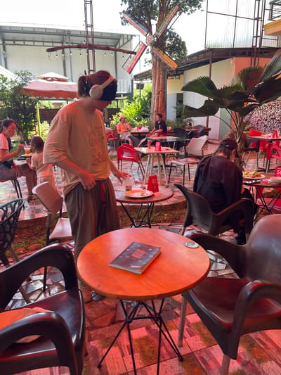 The image depicts an outdoor seating area of a cafe or restaurant, likely in Ia Hru, Vietnam, given the context. A young person with headphones on and a blindfold over their eyes is standing in the foreground, wearing a light-colored t-shirt and dark pants. They are holding a drink in their right hand. To the left, a woman in a pink shirt is seated, eating with a fork. A child in a light-colored outfit stands near her.

In the background, other patrons are visible seated at tables. One person in a black jacket is sitting at a table with a plate of food. The cafe features red chairs and tables, with some tables made of wood and metal. A large, ornate windmill structure is visible in the background, along with lush green vegetation. The ground is paved with patterned tiles. The overall atmosphere suggests a casual, open-air dining environment.