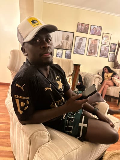 An indoor scene in Olkaria ward, Kenya, depicts three individuals in a residential setting. In the foreground, a dark-skinned male, approximately 20-30 years old, is seated casually on a light-colored, vertically striped armchair. He wears a light grey mesh baseball cap with a yellow "Gwethes" patch, a black short-sleeved t-shirt with gold Puma logos and a star design, and dark green patterned shorts. He holds a black smartphone in his left hand, looking upwards and to the right with his tongue slightly extended. To the right, in the middle ground, a dark-skinned female, also appearing 20-30 years old, is seated on a light-colored tufted sofa, wearing a yellow top and dark shorts, focused on a smartphone in her hands. A third individual's lower body is partially visible, seated on an adjacent sofa behind the main subject. The room features light yellow walls decorated with an arrangement of framed photographs, predominantly black and white portraits, and a tall wooden giraffe statue on the far right. The flooring visible on the left is wood. The lighting is artificial, creating a warm ambiance.