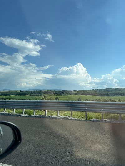 A scenic view of rolling green hills and a blue sky with scattered white clouds is captured from a roadside perspective. In the foreground, a dark asphalt road stretches across the lower portion of the image, with a portion of a car's side mirror visible on the left. A metal guardrail with vertical supports runs horizontally across the mid-ground, separating the road from the landscape beyond. The landscape consists of fields of varying shades of green, interspersed with patches of trees and denser forests on the distant hills. The sky is a vibrant blue, with dramatic white cumulus clouds, some of which appear to be building into storm clouds on the left side of the frame. The overall impression is of a rural or suburban environment on a clear, possibly humid day. The location is identified as Vakarel, Bulgaria.
