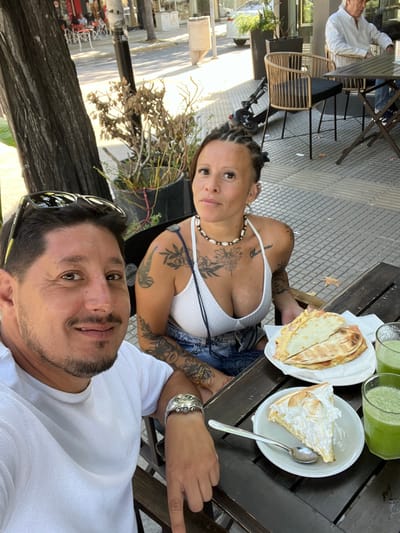 The image displays an outdoor cafe scene in City Bell, Argentina, during daylight. In the foreground, a man with short dark hair, a mustache, and a beard, wearing a white t-shirt and a silver watch, looks towards the camera with a slight smile. Sunglasses are perched on his head. Seated next to him is a woman with braided hair, numerous tattoos visible on her arms and chest, a white V-neck top, and denim shorts. She wears a shell necklace and multiple earrings and looks directly at the camera with a neutral expression. They are seated at a dark wooden slatted table. On the table, in front of the woman, is a white plate holding two folded food items, possibly quesadillas, with visible ham and cheese filling. In front of the man, a white plate contains a slice of meringue pie with a silver spoon. Two glasses of a green beverage are also present. In the background, an older man with white hair, wearing a white shirt, is seated alone at another outdoor table. The environment includes a checkered tiled sidewalk, potted plants (some appearing dry), a tree trunk, commercial buildings, and a parked scooter.