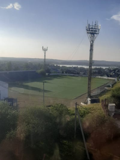 This is an aerial view of a stadium in the city of Shelekhov, Russia. The stadium features a green turf field with two white soccer goals. Spectator stands, partially covered in dark material, surround one side of the field. Two tall metal towers with lights are positioned on opposite sides of the stadium. In the background, residential buildings and a body of water are visible, with a hilly, tree-covered landscape beyond. The sky is a clear blue with a few wispy clouds. The image captures the stadium during daylight hours.