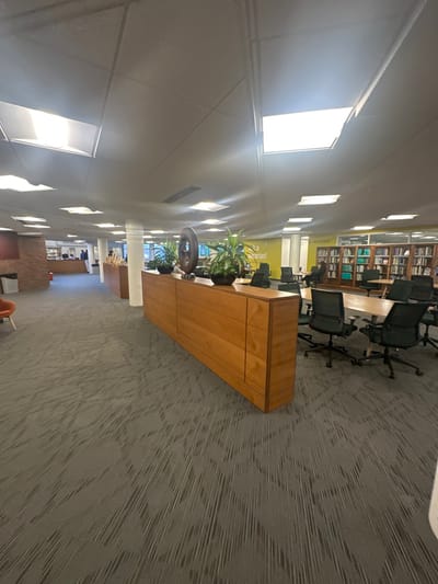 The image displays the interior of a library located in Wellesley, United States. The space features a dark gray patterned carpet and a white suspended ceiling with numerous square fluorescent light panels providing illumination.

In the left foreground, a section of an orange upholstered armchair is visible. To its right, a dark waste receptacle stands adjacent to a partial red brick wall element. Dominating the midground is a long, light-brown wooden cabinet, functioning as a partition. Placed atop this cabinet are two black planters containing green plants, separated by a dark, abstract, ring-shaped sculpture.

To the right of the wooden cabinet, a large, rectangular light-colored wooden table is surrounded by approximately six black office chairs with mesh backs and wheels. Further right, a partially obscured individual is seated at another table in the background.

In the left background, a service counter or reception desk is visible, behind which two to three individuals are present. A prominent yellow wall section in the center background displays the text "Ask a Librarian!" in dark letters. To the right, multiple tall wooden bookshelves, filled with books, line the wall, with a sign reading "Library Staff" mounted above them. Additional tables and chairs are scattered throughout the background area.