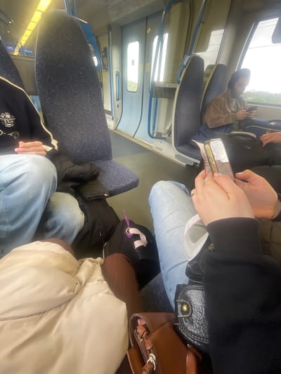 The image shows a first-person perspective of someone sitting on a train. The train's interior features blue upholstered seats with metal frames. Multiple passengers are visible, each occupied with their own activities. One person is looking at a phone, while the person taking the photo is holding a phone and a ticket. A black leather bag is beside the photographer. The setting is likely a commute, given the seating arrangement and the activity of looking at phones. The location is inferred as being on a train travelling in the city of St Albans, United Kingdom.