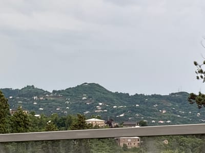 The image shows a landscape view of hills covered with trees and scattered houses in Chakvi, Georgia. The sky is overcast and gray. In the foreground, a gray railing is visible, with trees and buildings behind it. The hills are covered with dense green vegetation, and numerous small houses with red and white roofs are dotted across the slopes. The overall impression is of a rural or semi-rural setting in a mountainous region.