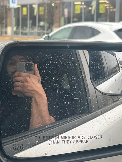 Captured in the Town of Cortlandt, United States of America, the image presents a view within a vehicle's side mirror. Rain droplets cover the mirror's surface. The reflection reveals a hand holding a smartphone. The mirror bears the inscription "OBJECTS IN MIRROR ARE CLOSER THAN THEY APPEAR". Outside the vehicle, a blurred building is visible. A white vehicle is partially seen in the mirror as well.