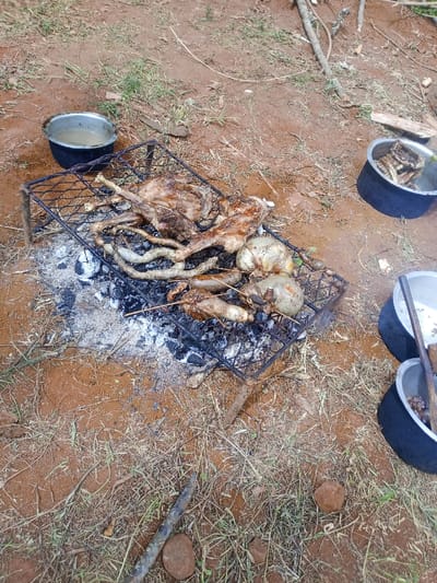 In Kiangochi, Kenya, an outdoor cooking scene is depicted on reddish-brown soil with sparse dry grass and scattered debris. Central to the image is a makeshift metal grill rack positioned over a bed of glowing charcoal embers and white ash. On the grill, various cuts of meat, some appearing to be lamb or goat, are undergoing roasting. Additionally, several long sections of intestines and other internal organs, some possibly stuffed or wrapped, are being cooked.
To the upper left of the grill, a silver metallic cooking pot (sufuria) with a black base contains a light-colored liquid, likely water or broth. To the upper right, another similar pot holds what appears to be cooked meat or bones. Towards the lower right, two more identical pots are visible; the upper one containing meat and a wooden stirring utensil, and the lower one partially visible, also containing solids. No individuals are present in the frame.
