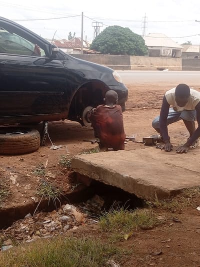 In Kwarau, Nigeria, a dark-colored sedan is elevated by a jack, its front passenger-side wheel removed and resting nearby on the dirt ground. A young boy, wearing a red garment, is crouched behind the vehicle, facing away from the camera, appearing to work on the exposed car parts. To the right, an adult male in a light-colored t-shirt and blue shorts is also crouched down, facing the same direction as the boy, actively engaged in manipulating something on the concrete slab in front of him, with tools or parts scattered around his hands. The environment is arid, with reddish-brown dirt and sparse patches of dry grass. In the foreground, a concrete structure with a visible drainage channel filled with debris is present. Beyond the car and the individuals, a paved road is visible, with buildings and vegetation in the background under an overcast sky.