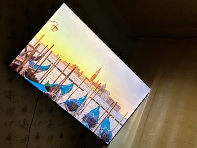 The image shows a television screen displaying a picturesque scene of gondolas docked in a canal, with a building and a tower in the background. The scene appears to be set in Venice, Italy, given the presence of gondolas and the architectural style of the buildings. The television is mounted on a wall covered with patterned wallpaper. The room is dimly lit, with the light from the television screen illuminating the immediate surroundings. There are no people visible in the image. The image does not provide any information about the city of Otradnaya, Russia.