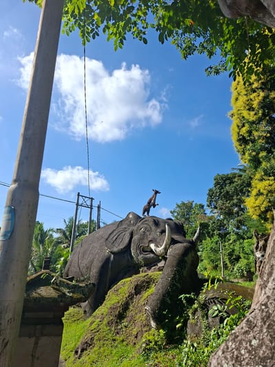 This image depicts a stone sculpture of an elephant, possibly a temple guardian or a monument, situated outdoors in Banjarangkan, Indonesia. The elephant sculpture is large, dark gray, and appears to be resting on a moss-covered hillock. It has prominent tusks and an open mouth. Perched on the elephant's head is a smaller, dark sculpture of an animal, possibly a deer or a goat, in a leaping pose. The background is dominated by a bright blue sky with scattered white clouds. Lush green foliage and trees surround the elephant, with some palm trees visible in the distance. A utility pole with electrical wires stands to the left of the elephant. The lighting suggests it is daytime, with sunlight casting shadows.