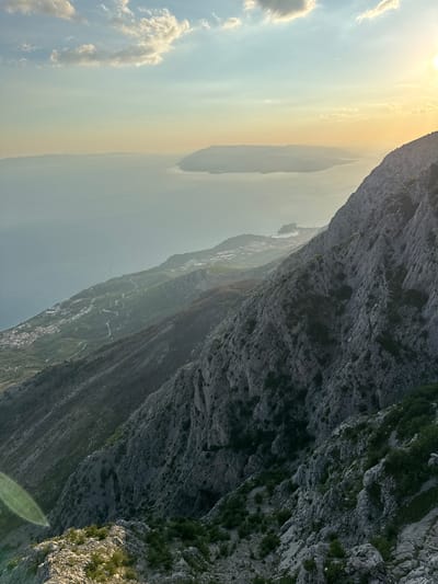 A high-angle, wide shot captures a coastal mountainous landscape at sunset in Općina Tučepi, Croatia. The foreground is dominated by a steep, rocky mountainside sparsely covered with green vegetation. Sunlight, originating from the upper right, casts a warm glow and creates lens flare in the lower left quadrant. Descending into the midground, the mountain slopes transition to a coastal plain featuring a winding road and a densely populated town with red-tiled roofs. This town is situated along the turquoise Adriatic Sea. In the background, a distant island and the hazy horizon are visible under a sky filled with soft clouds and the fading sun.