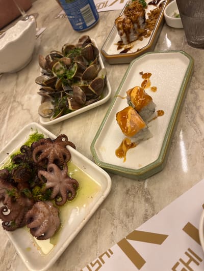The image shows a table setting with multiple plates of food. There are three distinct dishes visible. On the left is a plate with what appears to be clams. Adjacent to it, there's a plate with grilled octopus, topped with a green herb sauce. A third plate contains pieces of sushi with a orange-brown glaze. The table surface is a light marble-patterned texture, partially covered with a paper or menu. The environment suggests a restaurant setting. The restaurant's name appears to have the word "Sushi" in it. There are no people visible in the image. The location could be Torreforta, Spain, based on the request, but no direct evidence in the image confirms this location.