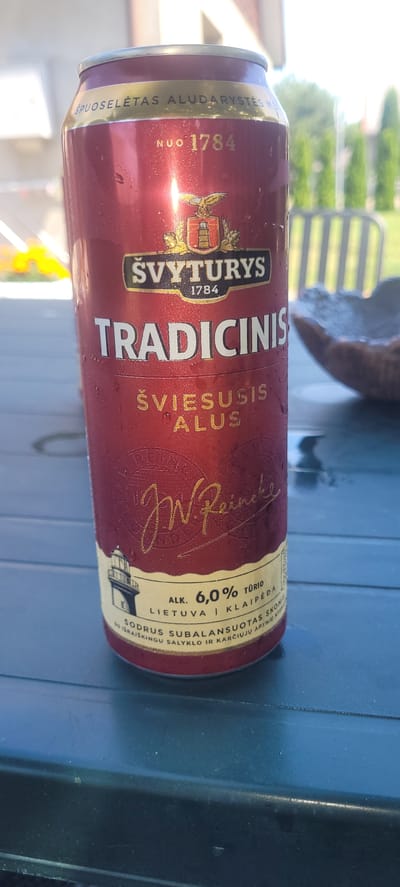 A close-up shot captures a red beer can with condensation drops, placed on a dark blue, textured surface, likely a table. The can features a gold and white label with embossed text and a crest. Prominently displayed are the words "TRADICINIS" and "ŠVYRURYS," along with a year, "1784." Below these, the text "ŠVIESIUSIS ALUS" appears. Further down, a lighthouse illustration is visible, and text indicates "ALK. 6.0% TŪRIO" and "LIETUVA | KLAIPĖDA." The background is softly blurred, showing a glimpse of green foliage and a white structure, suggesting an outdoor setting. The overall impression is that of a beverage advertisement or product shot.
