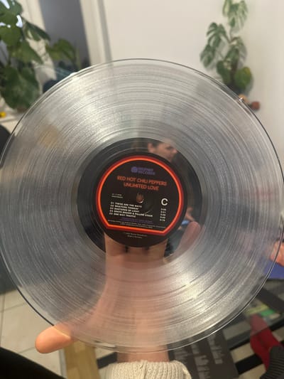 The image shows a clear vinyl record, a special edition of Red Hot Chili Peppers' "Unlimited Love" album. The label displays the Warner Records logo along with the album name and the band. Side C of the record lists the tracks: "These Are the Ways," "Whatchu Thinkin'," "Bastards of Light," "White Braids & Pillow Chair," and "One Way Traffic." The vinyl is held in someone's hand, with a blurred background suggesting an indoor setting.