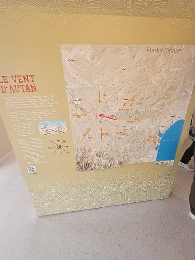 The image displays an indoor informational exhibit panel, likely within a public or educational facility in Avignonet-Lauragais, France. The panel, vertically oriented and light beige with subtle contour patterns, details "LE VENT D'AUTAN" (The Autan Wind). The left side presents the title in red capital letters, followed by French explanatory text, two small illustrative diagrams (one appearing to be wind turbines, the other a wind rose), and a QR code. The right side features a raised topographical map of a region in France, including the Massif Central, with departmental names like Tarn, Aveyron, Haute Garonne, and Aude. Orange arrows on the map illustrate wind patterns converging towards a central point labeled "LAURAGAIS" with a red arrow, and a blue area represents the Mediterranean Sea. The display rests on a light grey, speckled floor. A person's lower right leg and foot, clad in a green shoe with a white sole, are partially visible on the far right, indicating a viewer interacting with the exhibit.