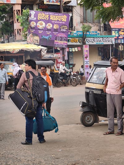 The image depicts a street scene in Datala, India, focusing on the front of several shops and a street corner. In the foreground, a man wearing a pink striped shirt and grey pants stands by an auto-rickshaw, gesturing with his hands. To his left, another man, dressed in a brown shirt and blue jeans, carries a large black backpack and a blue duffel bag. Several other individuals are visible in the background, including a woman wearing a white headscarf near a yellow auto-rickshaw, and men standing or riding motorcycles. Above the shops, a large purple banner advertises "Hotel Bhayashri" with images of food. Shops on the right include "Shubham Generic Medicine" and "Shubham Pan Mandir". The environment appears to be a busy marketplace with pedestrians, vehicles, and commercial establishments. The overall lighting suggests it is daytime, possibly late morning or afternoon.