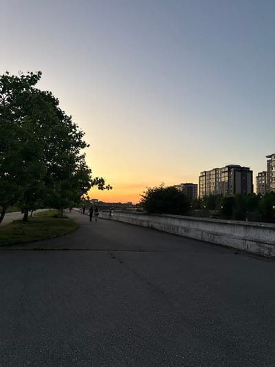 The image depicts a sunset scene along a paved walkway in Khachta, Russia. A large tree with dense green foliage is situated on the left side of the walkway, with grass and smaller trees behind it. Several people are visible walking along the path, some appearing to be couples. On the right side of the walkway, a concrete barrier separates it from a body of water, beyond which are several tall, multi-story apartment buildings. The sky transitions from a pale blue at the top to a vibrant orange and yellow near the horizon, indicating the setting sun. The overall atmosphere is calm and serene, suggesting an evening stroll.