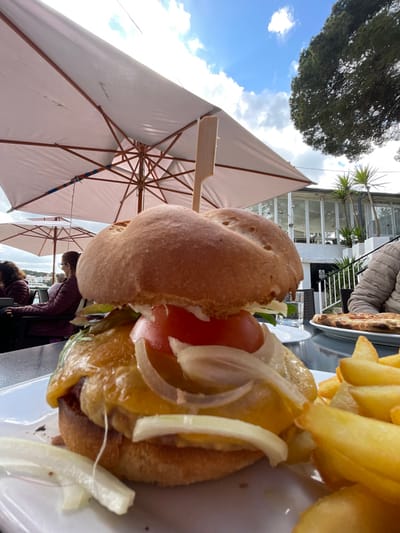 The image depicts an outdoor dining scene at a restaurant patio in Porto Cristo, Spain. In the immediate foreground, a large hamburger and a portion of thick-cut french fries are presented on a white rectangular plate, resting on a dark-colored table. The hamburger features a toasted, seeded bun, a meat patty covered with melted yellow-orange cheese, sliced white raw onion rings, a thick red tomato slice, and green leaf lettuce. A slender wooden skewer is inserted vertically through the center of the burger. The french fries are golden-brown.
Overhead, multiple large white patio umbrellas with visible wooden support structures provide shade. To the left, two individuals are seated at a separate table. The closest person wears a dark maroon jacket and is seen from behind, looking towards a body of water with white buildings visible on the distant shore, indicative of a harbor or bay. To the right, a third individual, partially visible, is seated at another table with a round pizza on a white plate in front of them.
The background features a multi-story white building with numerous large glass windows and balconies, consistent with a restaurant or hotel structure. Green foliage, including tall trees and smaller palm-like plants, surrounds the building. The sky is blue with scattered white clouds, suggesting a sunny day.