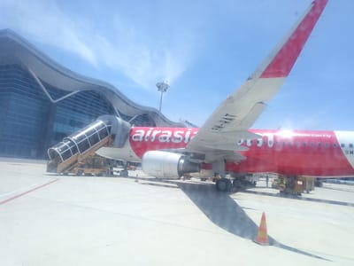 An AirAsia airplane is parked on the tarmac at Cam Ranh International Airport (CXR) in Cam Ranh City, Vietnam. The airplane, a red Airbus with white lettering that reads "airasia Everyone Can Fly," has its passenger stairs attached to the front left door. The airport terminal building, with its distinctive curved, modern architecture and glass facade, is visible in the background. The sky is bright blue with scattered white clouds. Several ground vehicles, including a baggage tug and a stair truck, are positioned around the aircraft. A few airport personnel can be seen near the plane and the vehicles. The ground is a light-colored concrete tarmac, marked with yellow and red lines. A bright orange and yellow traffic cone is positioned in the foreground. The scene suggests the aircraft is either preparing for departure or has recently arrived.