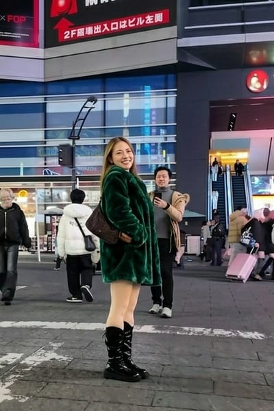 The image depicts an urban pedestrian area at night in Gifu, Japan. A light-skinned female individual with long light-brown hair stands center-left, smiling towards the camera. She wears a dark green faux fur coat, a short light-colored garment exposing her upper thighs, and tall, shiny black boots. A dark-patterned handbag is slung over her right shoulder. Behind her, to the right, a male individual of East Asian descent, wearing a beige jacket over a grey turtleneck, holds a smartphone up, appearing to film the female subject.

The background features modern multi-story buildings with glass facades and illuminated signage. A prominent digital sign at the top center displays Japanese text, including "2F階増出口はビル左側" (2nd-floor extension exit is on the left side of the building), along with a red circular graphic. To the far left, an older male in a black jacket walks away from the camera. Another individual in a white hooded puffer jacket and black pants walks away in the midground-left. To the right, a group of people is visible near escalators leading into a building, with one person pulling a pink rolling suitcase. The pavement is paved with light-colored blocks and shows patches of reflectivity, possibly from moisture. Artificial lighting from the surrounding buildings and signs illuminates the scene.