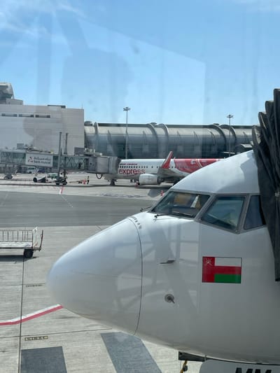The scene depicts an airport tarmac at Muscat International Airport in Seeb Province, Oman, observed through a window under a clear blue sky. In the immediate foreground, the white nose and cockpit section of an aircraft, likely connected to a jet bridge on the right, features a decal of the Omani flag below the cockpit windows. In the midground, an Air India Express aircraft, predominantly white with a red tail and engine housing, is parked at a gate with a jet bridge attached to its forward door. The jet bridge displays "بنك مسقط bank muscat" branding. An airport ground staff member, identifiable by a yellow high-visibility vest, is visible near the connection point of the jet bridge to the Air India Express aircraft. Various ground support equipment, including a metal luggage cart, are present on the tarmac, which features "14L 110" markings. The background shows part of the airport terminal building with a distinctive arched, metallic roof structure and several tall light poles.