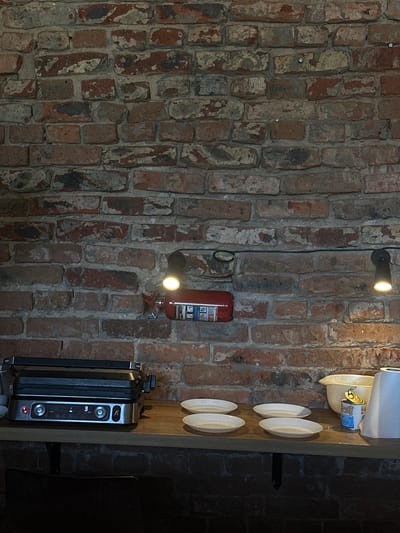 A close-up shot captures a section of a rustic brick wall, with a wooden shelf mounted against it. On the shelf, a silver panini press is prominently displayed on the left. To its right, a stack of four light-colored plates sits arranged in two rows. Further to the right, a pale yellow mixing bowl is visible, next to a can of food and a roll of paper towels on the far right. Above the shelf, a red fire extinguisher is mounted on the wall, illuminated by a light fixture. Another light fixture is positioned to the right of the fire extinguisher, casting a warm glow. The brickwork itself is varied in color, featuring shades of red, brown, and gray, with visible mortar. The overall scene suggests a functional kitchen or dining area within a rustic setting.