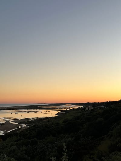 The image depicts a twilight or sunset scene, possibly at the mouth of the Guadiana River bordering Vila Real de Santo António, Portugal. The sky transitions from a light blue-grey to a warm orange. The river or estuary is visible on the right, with patches of exposed sand and boats dotting the water's surface. Lush, dark greenery covers the riverbank, interspersed with a few buildings.