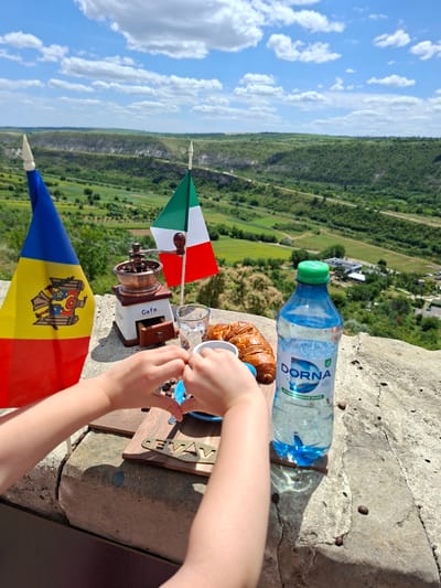 In Trebujeni, Moldova, on a stone ledge overlooking a verdant valley under a partly cloudy sky, a scene is set for a rustic breakfast. Two small flags, one the national flag of Moldova and the other the flag of Italy, stand upright. In front of them, a vintage-style manual coffee grinder sits next to a clear glass of water and a croissant. A bottle of "DORNA" mineral water with a green cap is positioned to the right. Below these items, a child's hands are actively arranging coffee beans on a small wooden board inscribed with the word "CEVAV". The background reveals a vast landscape with rolling green hills, a winding dirt road, and scattered buildings. The lighting suggests a bright, sunny day.